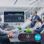 A business team meets in a modern conference room as a presenter points to a screen showing financial charts and market data.
